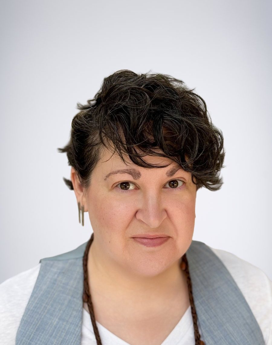 Kelly Rogers, with short dark curly hair, a white tee and a chambray vest, giving a slight knowing smile and a subtly raised eyebrow.